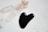 A smooth, black, heart-shaped stone is placed on a light marble surface beside several clear quartz crystals. In the background, a small glass bottle with an orange-toned liquid is visible, partially out of focus.