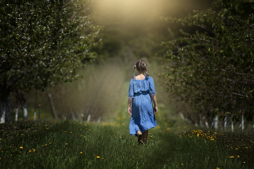 girl in blue dress walking on grass field by trees at daytime