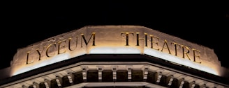 An illuminated sign atop a building displaying the words 'Lyceum Theatre' in large golden letters against a dark background. The architectural details below the sign suggest a classical style, with ornate moldings and decorative elements. The lighting highlights the textures and engravings on the facade.