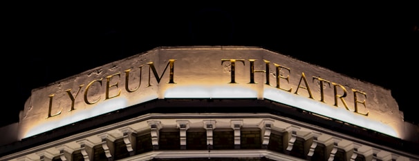 An illuminated sign atop a building displaying the words 'Lyceum Theatre' in large golden letters against a dark background. The architectural details below the sign suggest a classical style, with ornate moldings and decorative elements. The lighting highlights the textures and engravings on the facade.