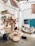 A cozy, rustic room with wooden furniture and decor elements. There is a wooden bookshelf filled with books and plants, a hanging hammock chair, and a pallet sofa with cushions. The room features various natural elements, including woven baskets and a jute rug. A blue door and a map on the wall add color to the bright, airy space.