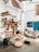 A cozy, rustic room with wooden furniture and decor elements. There is a wooden bookshelf filled with books and plants, a hanging hammock chair, and a pallet sofa with cushions. The room features various natural elements, including woven baskets and a jute rug. A blue door and a map on the wall add color to the bright, airy space.