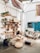 A cozy, rustic room with wooden furniture and decor elements. There is a wooden bookshelf filled with books and plants, a hanging hammock chair, and a pallet sofa with cushions. The room features various natural elements, including woven baskets and a jute rug. A blue door and a map on the wall add color to the bright, airy space.