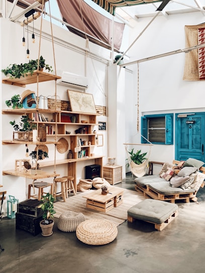 A cozy, rustic room with wooden furniture and decor elements. There is a wooden bookshelf filled with books and plants, a hanging hammock chair, and a pallet sofa with cushions. The room features various natural elements, including woven baskets and a jute rug. A blue door and a map on the wall add color to the bright, airy space.