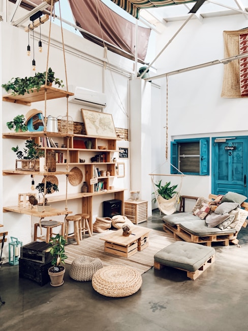 A cozy, rustic room with wooden furniture and decor elements. There is a wooden bookshelf filled with books and plants, a hanging hammock chair, and a pallet sofa with cushions. The room features various natural elements, including woven baskets and a jute rug. A blue door and a map on the wall add color to the bright, airy space.
