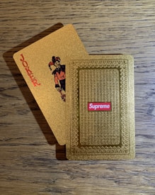 A pair of golden playing cards on a wooden table, one face displaying a joker with the word 'JOKER' printed vertically and the other showing an ornate pattern with a 'Supreme' logo in red.