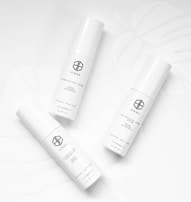 Three white cylindrical skincare product bottles are arranged on a white background with subtle leaf patterns. Each bottle has minimalist black text and logos, conveying a clean and modern aesthetic.
