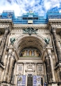 An architectural structure with classical elements such as tall columns and elaborate archways. A detailed mural is situated above the door, depicting several human figures in historical attire. The building is under some form of restoration or construction, as indicated by the blue scaffolding present on the upper portions. The sky in the background shows a mix of clouds and blue sky.