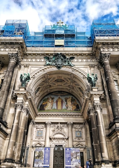 An architectural structure with classical elements such as tall columns and elaborate archways. A detailed mural is situated above the door, depicting several human figures in historical attire. The building is under some form of restoration or construction, as indicated by the blue scaffolding present on the upper portions. The sky in the background shows a mix of clouds and blue sky.