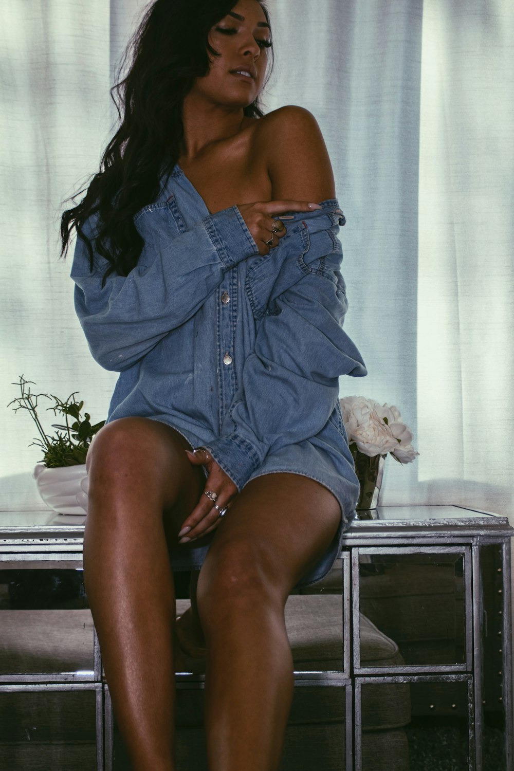 woman wearing blue denim jacket sitting on end table near window curtain