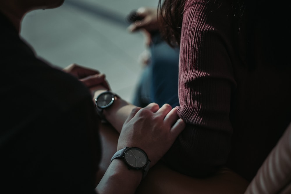 person wearing round silver-colored analog watch with leather band sitting near another person