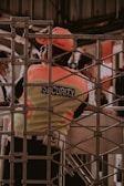 A security guard is seen wearing a fluorescent vest and cap, leaning against a turnstile gate. The setting appears to be in an indoor or enclosed area with a metal grid structure in the foreground. Other people are partially visible in the background, engaged in different activities.