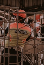 A security guard is seen wearing a fluorescent vest and cap, leaning against a turnstile gate. The setting appears to be in an indoor or enclosed area with a metal grid structure in the foreground. Other people are partially visible in the background, engaged in different activities.