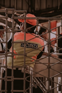 A security guard is seen wearing a fluorescent vest and cap, leaning against a turnstile gate. The setting appears to be in an indoor or enclosed area with a metal grid structure in the foreground. Other people are partially visible in the background, engaged in different activities.