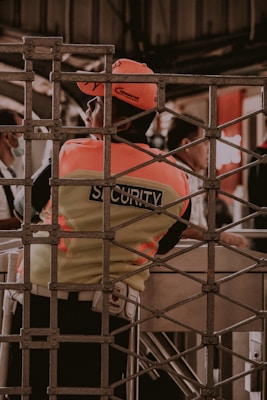 A security guard is seen wearing a fluorescent vest and cap, leaning against a turnstile gate. The setting appears to be in an indoor or enclosed area with a metal grid structure in the foreground. Other people are partially visible in the background, engaged in different activities.