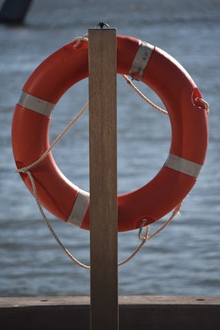 A bright orange lifebuoy is attached to a wooden post, positioned against the background of a calm water body. The lifebuoy is secured with white ropes, and the sunlight casts subtle shadows on it.