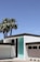 A modern house with a minimalistic design featuring clean white walls and a flat roof. A prominent turquoise door provides a striking accent to the building. Two tall palm trees tower beside the structure, under a clear blue sky.