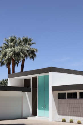 A modern house with a minimalistic design featuring clean white walls and a flat roof. A prominent turquoise door provides a striking accent to the building. Two tall palm trees tower beside the structure, under a clear blue sky.