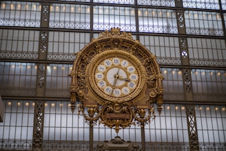 A large, ornate clock with Roman numerals is mounted on an elaborate metal framework. The background consists of a grid-like pattern of large windows, allowing natural light to illuminate the clock.