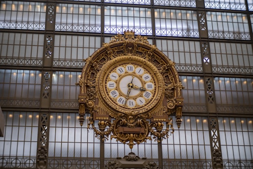 A large, ornate clock with Roman numerals is mounted on an elaborate metal framework. The background consists of a grid-like pattern of large windows, allowing natural light to illuminate the clock.