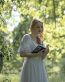 A person with red hair is standing in a lush, green outdoor setting, holding a small book. The soft focus background creates a dreamy atmosphere with light filtering through the leaves, casting a warm glow.