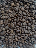 A close-up view of a large pile of roasted coffee beans. The beans are uniformly dark brown, with visible creases and a shiny surface indicating freshness and oil.