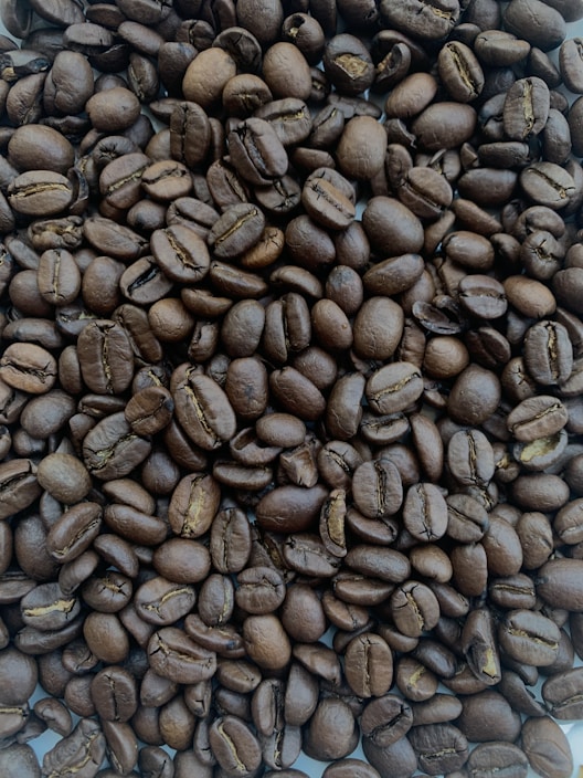 A close-up view of a large pile of roasted coffee beans. The beans are uniformly dark brown, with visible creases and a shiny surface indicating freshness and oil.