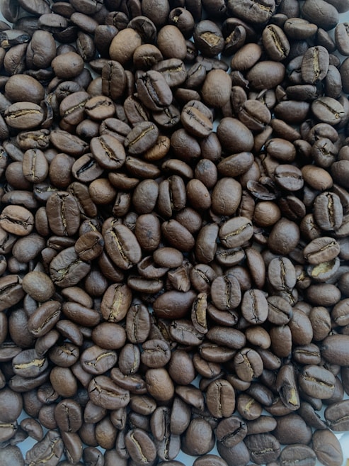 A close-up view of a large pile of roasted coffee beans. The beans are uniformly dark brown, with visible creases and a shiny surface indicating freshness and oil.