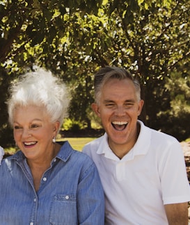 An elderly woman with white hair and a middle-aged man with gray hair are sitting together outdoors. They are both smiling and appear joyful. The woman is wearing a denim shirt, and the man is in a white polo shirt. They are surrounded by leafy green trees, suggesting a park or garden setting.