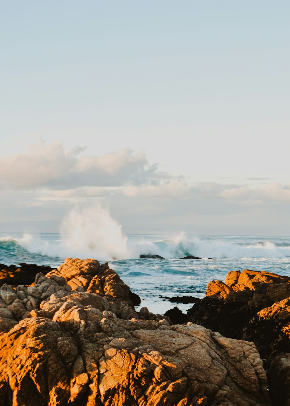 formações rochosas marrons que visualizam o corpo de água com grandes ondas sob o céu branco e azul durante o dia