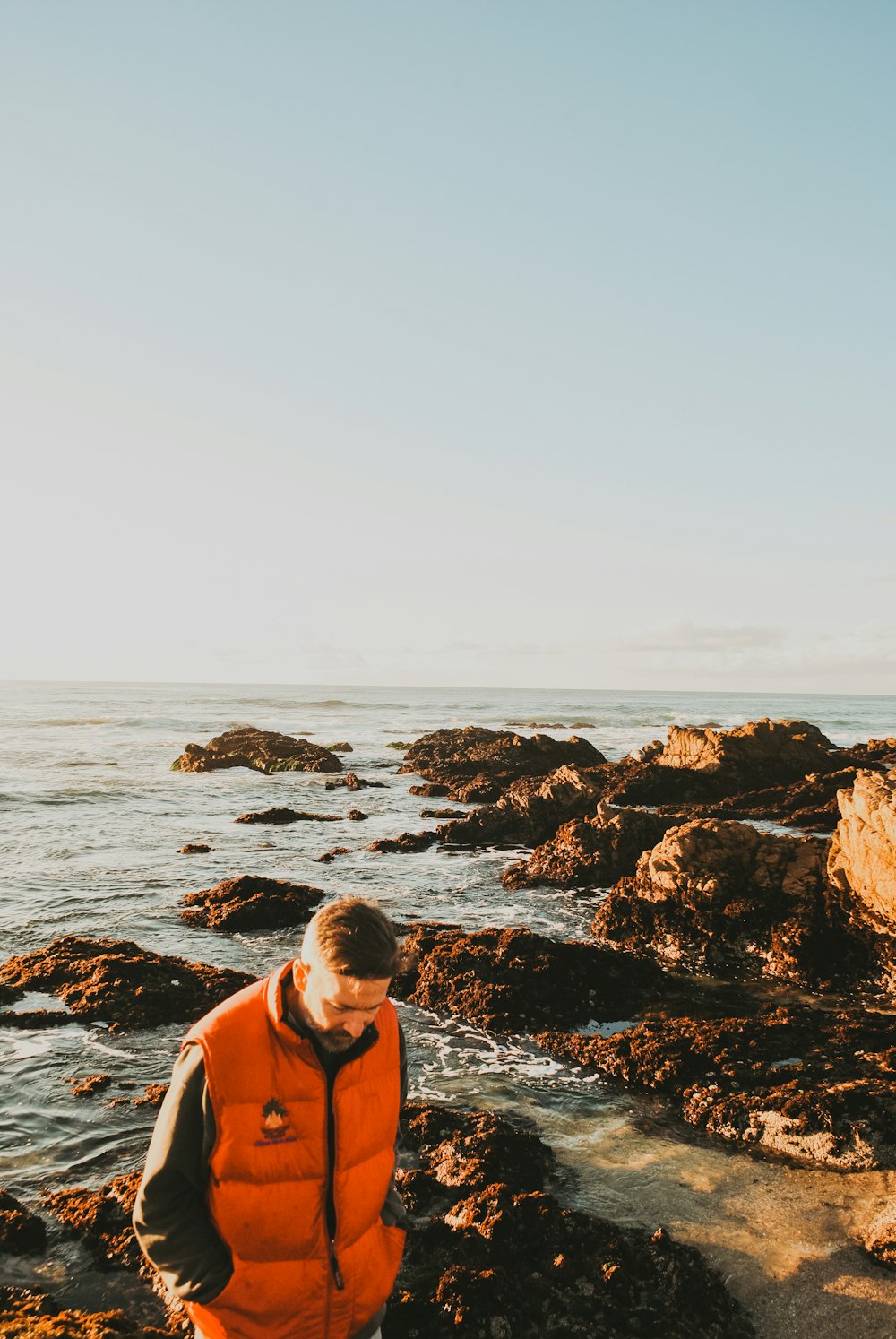 Homme portant une veste à bulles orange et noire debout sur un rocher tout en regardant vers le bas en regardant le plan d’eau calme pendant la journée