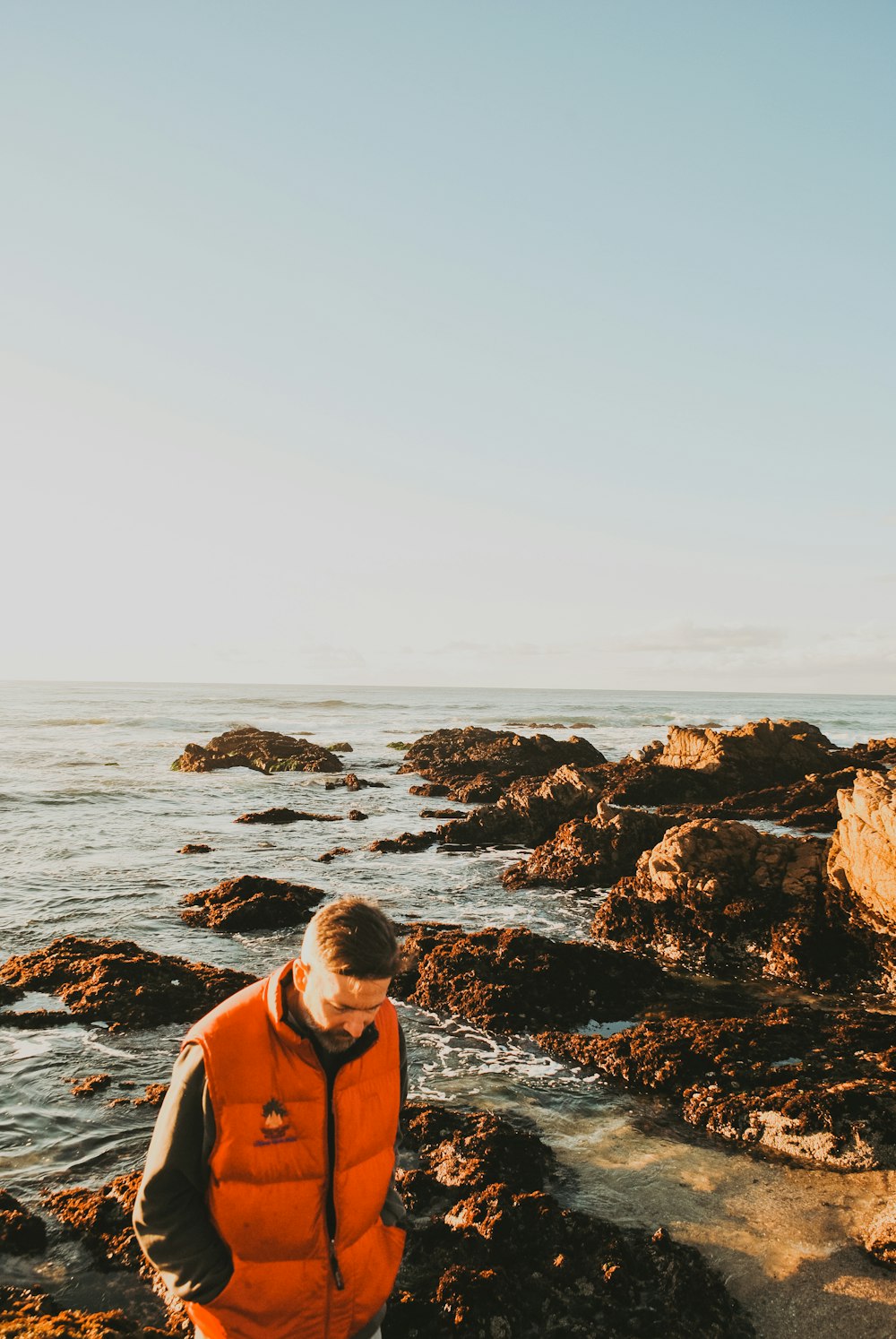 man wearing orange and black bubble jacket standing on rock while looking down viewing calm body of water during daytime