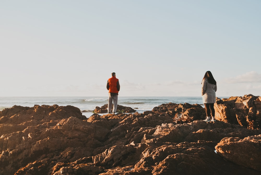 two person standing on brown rocks viewing body of water during daytime