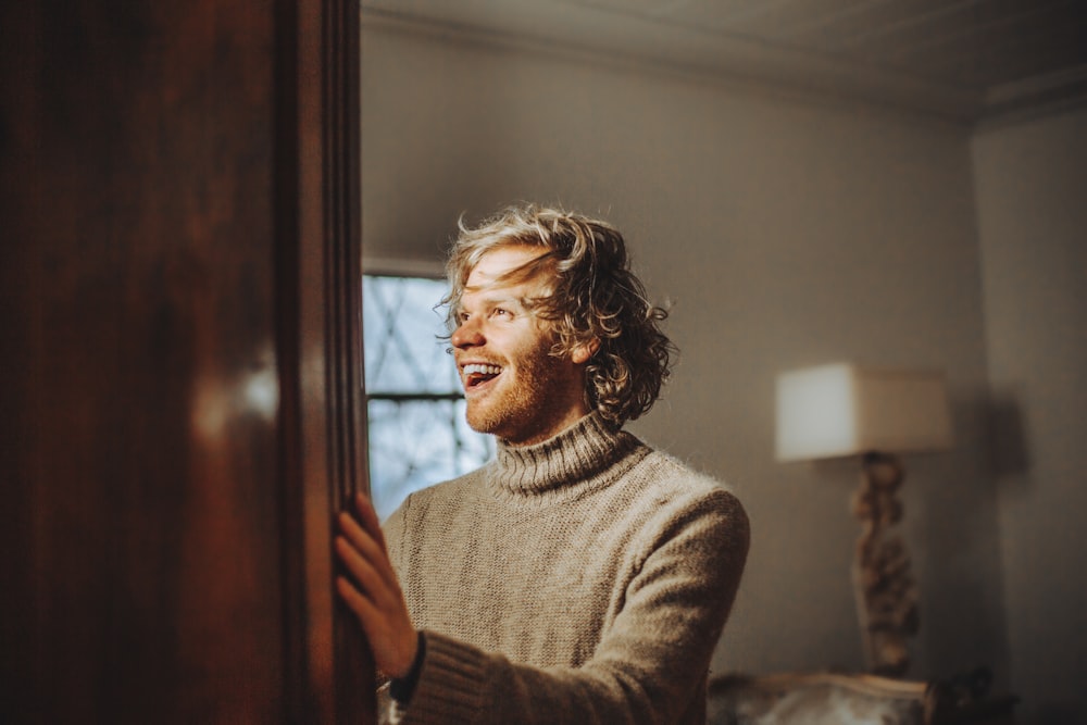 hombre con suéter marrón de pie y sonriendo cerca de la pared