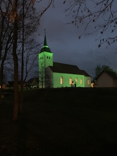 A church with illuminated green lighting stands in a dark, outdoor setting. The church features a tall steeple with a cross at the top. Trees with bare branches frame the scene, contributing to the somber mood. Soft light glows through the church’s windows, suggesting it is evening or night.