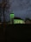 A church with illuminated green lighting stands in a dark, outdoor setting. The church features a tall steeple with a cross at the top. Trees with bare branches frame the scene, contributing to the somber mood. Soft light glows through the church’s windows, suggesting it is evening or night.