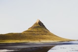 A large, steep mountain rises sharply from a flat landscape with minimal vegetation, under a clear sky. The lower parts of the mountain are covered in shades of brown and green, while the summit is more rugged and rocky. The foreground features a mix of dark sand and shallow water, perhaps a bay or tidal flat.
