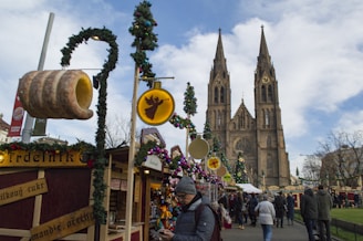A festive market scene with people walking among stalls decorated with Christmas lights and wreaths. A large Gothic-style cathedral towers in the background. Various goods are displayed, including ornaments and a large sign of a traditional baked treat.