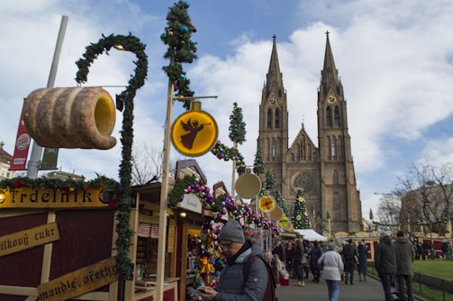 A festive market scene with people walking among stalls decorated with Christmas lights and wreaths. A large Gothic-style cathedral towers in the background. Various goods are displayed, including ornaments and a large sign of a traditional baked treat.