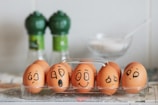 A row of brown eggs displayed in a transparent plastic tray, each egg has a different expressive face drawn on it with black marker. The background features two green-capped spice bottles and a clear dish containing a white substance, possibly sugar or salt.