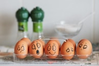 A row of brown eggs displayed in a transparent plastic tray, each egg has a different expressive face drawn on it with black marker. The background features two green-capped spice bottles and a clear dish containing a white substance, possibly sugar or salt.