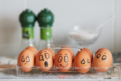 A row of brown eggs displayed in a transparent plastic tray, each egg has a different expressive face drawn on it with black marker. The background features two green-capped spice bottles and a clear dish containing a white substance, possibly sugar or salt.