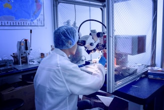 A person in a laboratory setting works at a microscope station, wearing a white lab coat, blue hair net, and blue gloves. The environment is sterile, with scientific equipment organized around the station. A world map is visible on the wall, and a netted barrier separates sections of the lab.