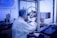 A person in a laboratory setting works at a microscope station, wearing a white lab coat, blue hair net, and blue gloves. The environment is sterile, with scientific equipment organized around the station. A world map is visible on the wall, and a netted barrier separates sections of the lab.