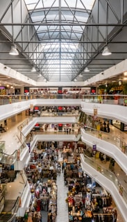 A spacious indoor shopping mall with multiple floors featuring a variety of shops and stalls. The architecture includes a large glass skylight allowing natural light to illuminate the central area. There are escalators connecting the different levels, and several people can be seen walking around or browsing the stores.