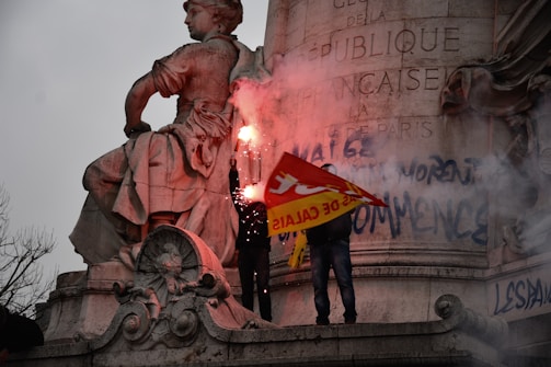 A large stone statue featuring a seated figure, possibly a historical or symbolic representation, is in the foreground. Two individuals stand near it, holding a red and yellow flag with French text and burning flares that emit bright light and smoke. Graffiti is visible on the stone surface behind them. The scene appears to be in a public or commemorative space, indicated by the engraved text in French on the stone.