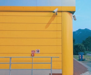 A vibrant yellow wall forms the backdrop, featuring two security cameras positioned at the top corner. A small red sign with a hand symbol and another with text can be seen below. In the foreground, a light gray railing runs horizontally. Beyond the wall, there is a view of mountains under a clear blue sky, and a trash bin is located near the path beside the building.