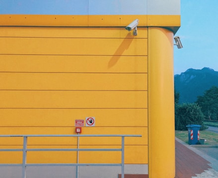 A vibrant yellow wall forms the backdrop, featuring two security cameras positioned at the top corner. A small red sign with a hand symbol and another with text can be seen below. In the foreground, a light gray railing runs horizontally. Beyond the wall, there is a view of mountains under a clear blue sky, and a trash bin is located near the path beside the building.