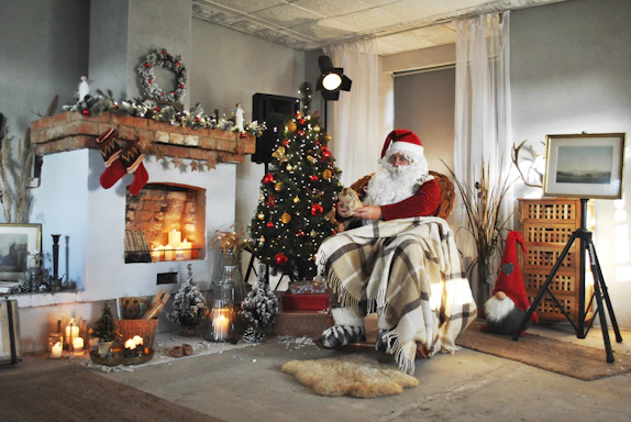 A cozy living room decorated for Christmas with a warm fireplace and a Santa hat on a chair.