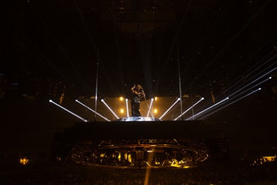 A performer stands elevated on a circular stage, surrounded by bright spotlights and beams of light radiating outward. The setting appears to be a concert or live performance with an audience dimly visible below.